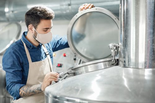 Fermentation and beverage production in modern plant. Young attractive male worker with tattoos in apron and protective mask opens lid on equipment in interior of brewery with kettles, profile